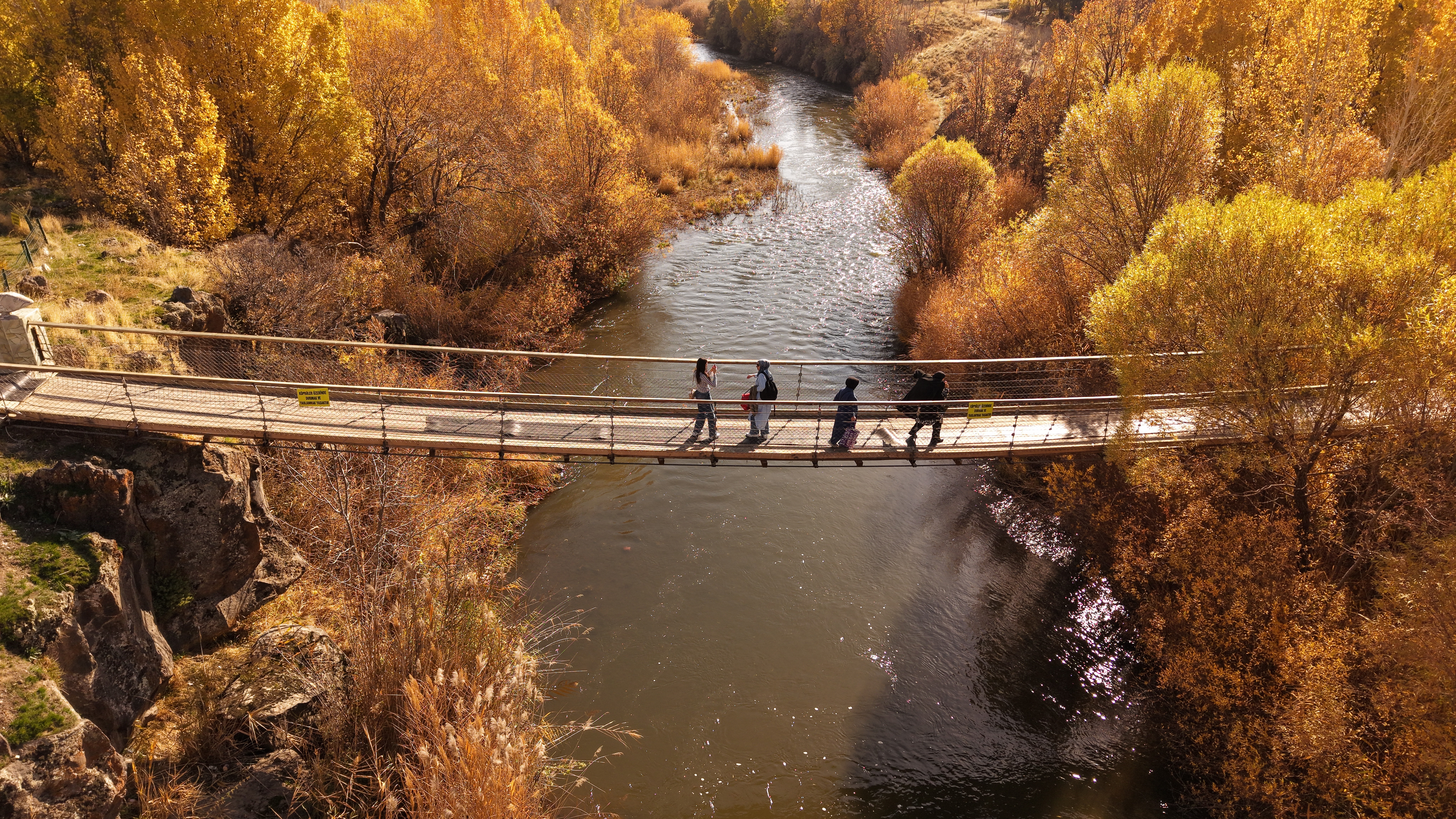 Several people walk on a foot bridge over a river, surrounded by trees in yellow fall colors.