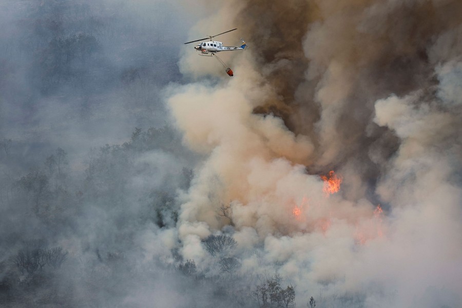 A helicopter flies with a water bucket above a large forest fire.