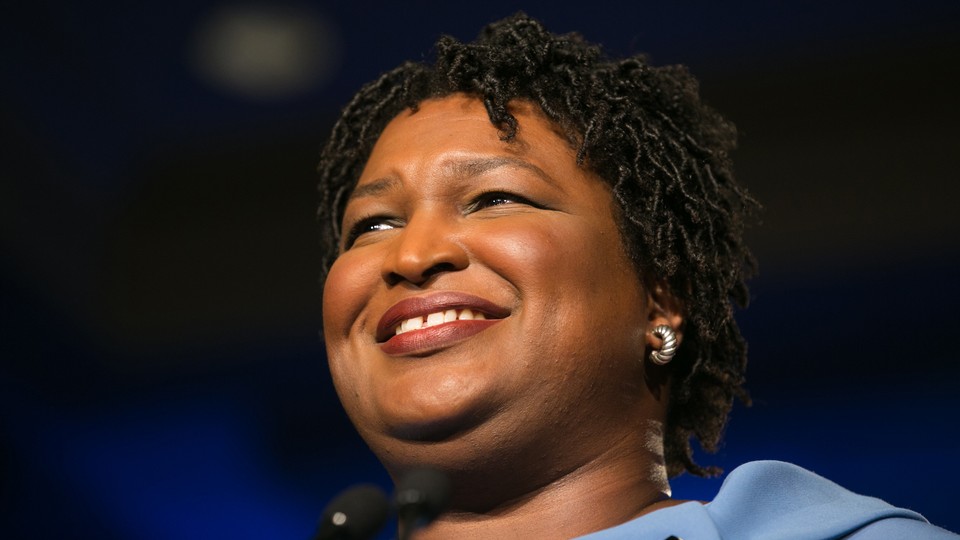 Stacey Abrams looking into the distance. She is smiling and wearing a blue dress.