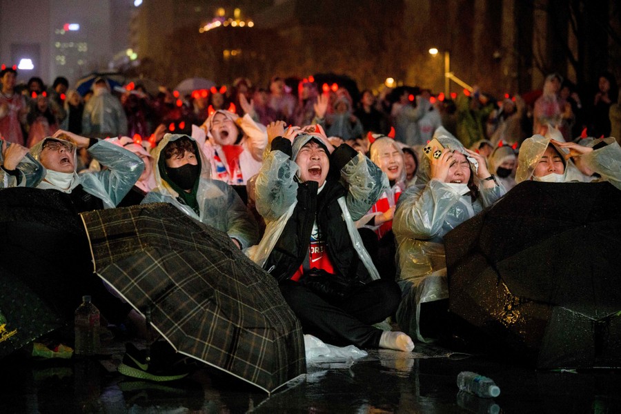 Fans sit on the ground, watching a game, covering their heads and making pained reactions.