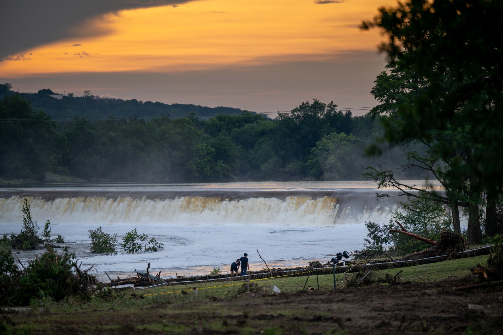 Two people stand beside a rushing river that has recently flooded.
