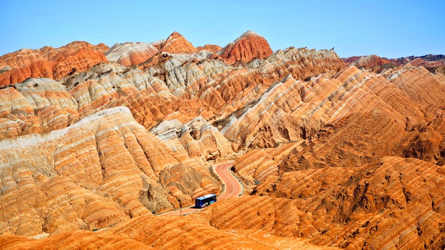 An elevated view of a bus driving on a winding road through layered rocky hills
