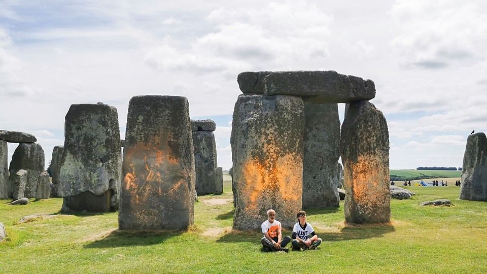 A photo of two protesters sitting in front of Stonehenge, which has orange spray paint on it