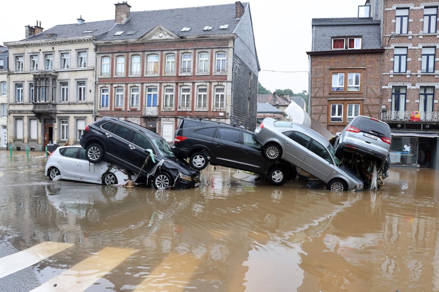 At least five cars are seen piled on each other, rear ends resting on hoods, among receding floodwater in a street.