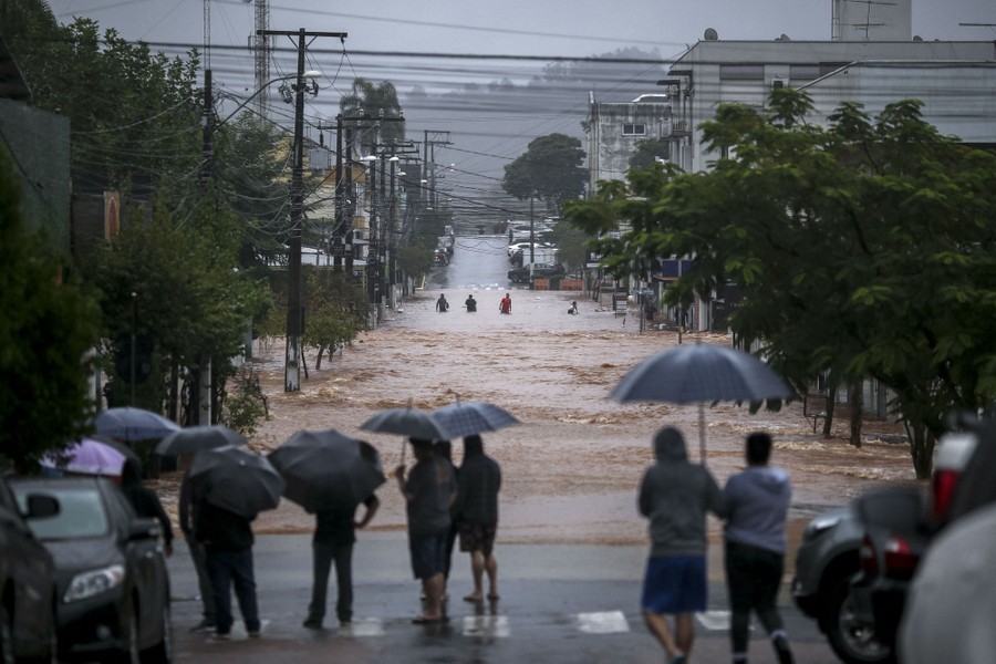 People stand on a street holding umbrellas while looking toward a flooded road.