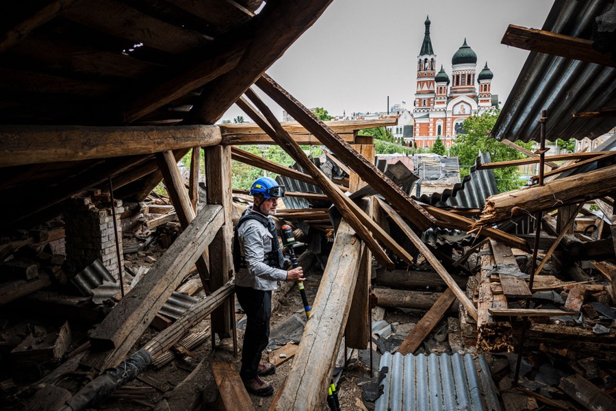 A person carrying a handheld scanner stands inside a damaged building, with other buildings visible through huge holes in the roof.
