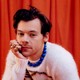 Harry Styles in a white sweater, blue chunky necklace, against orange-red curtain backdrop
