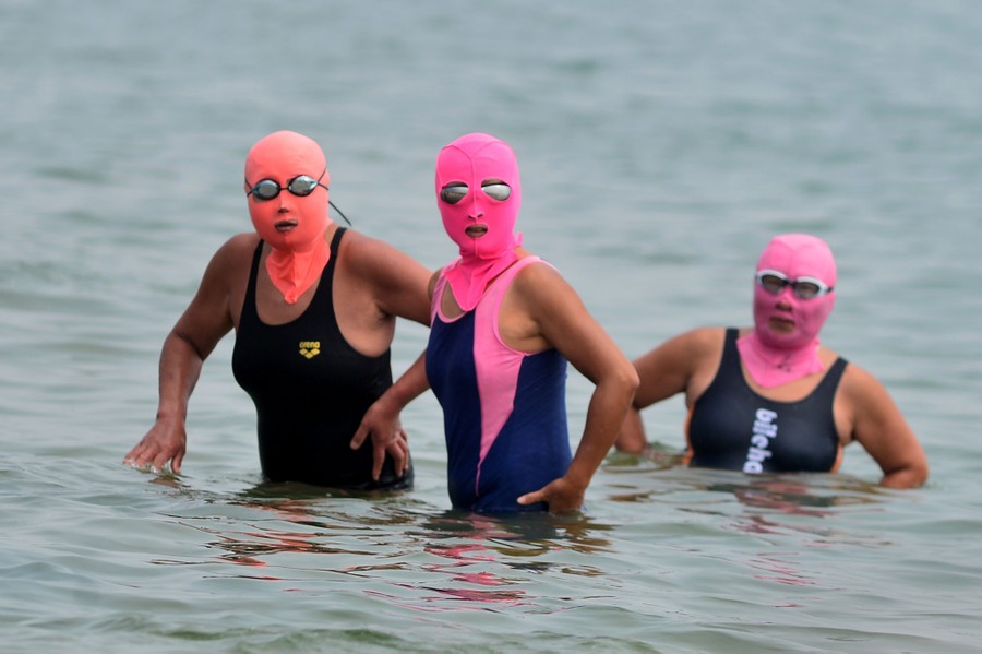 Three people in waist-deep water wear bathing suits and head coverings that cover their face as well.
