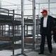 Photograph of Donald Trump in MAGA hat looking at rows of empty cots behind chain-link bars