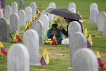 A child sits under an umbrella with an adult in a cemetery. Colombian flags are in the ground next to each headstone.