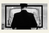 A black-and-white image of a man facing the front of a car
