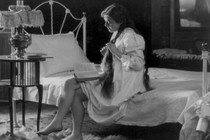 A young woman reads at the corner of her bed as she braids her hair.