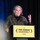 Steve Bannon gestures as he speaks at a podium with yellow text that says "The Pro-Human Assembly 2026"