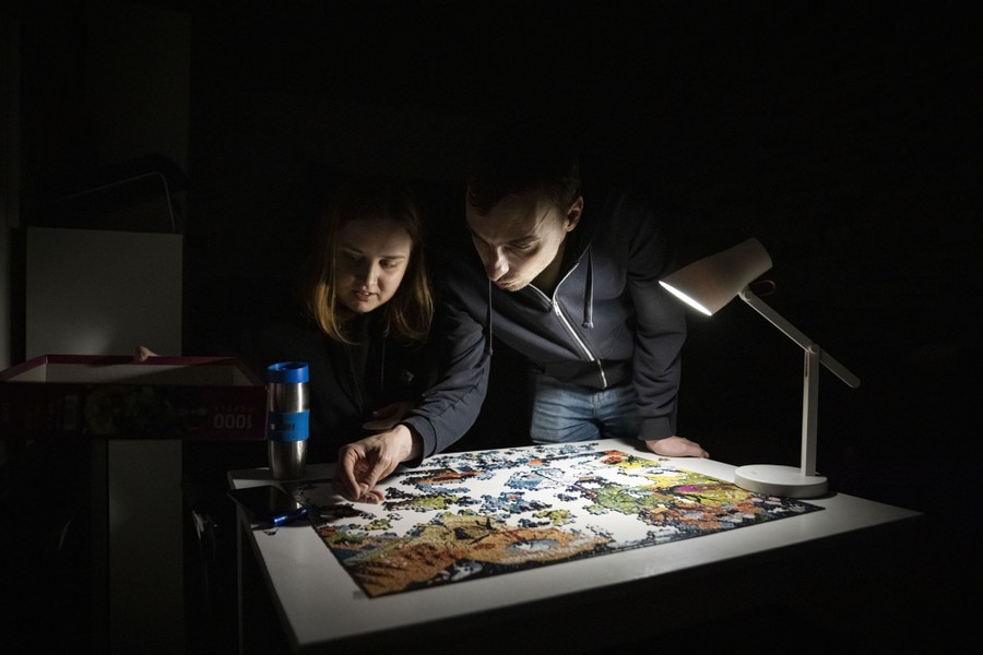Two people work on a jigsaw puzzle lit by a small lamp, in a dark apartment.