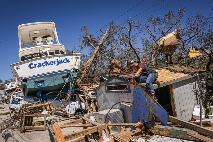 A person works on clearing piles of storm debris, with a grounded boat in the background.