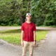 Color photograph of Rob Sand, wearing a red T-shirt and khaki pants with tennis shoes and a white ball cap, standing in front a tree.