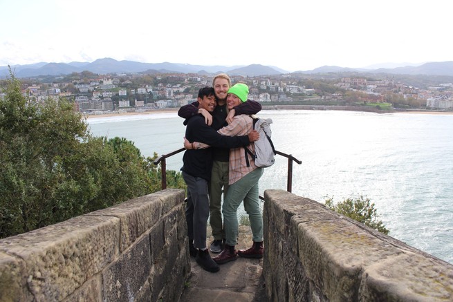 On a stone bridge overlooking a body of water, two men and a woman share a group hug. There are mountains and a town in the distance on the other side of the water.