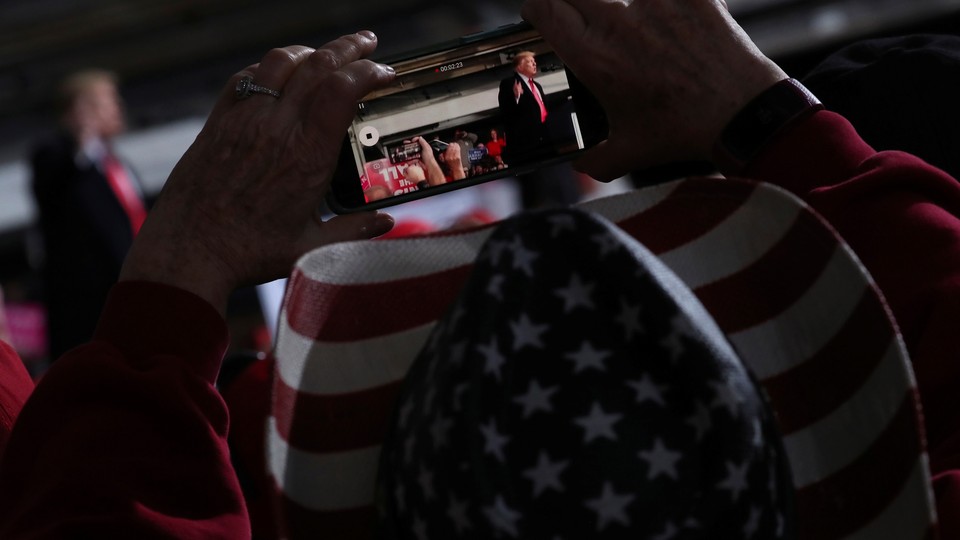 A voter in a hat patterned with an American flag films Donald Trump with a handheld phone.