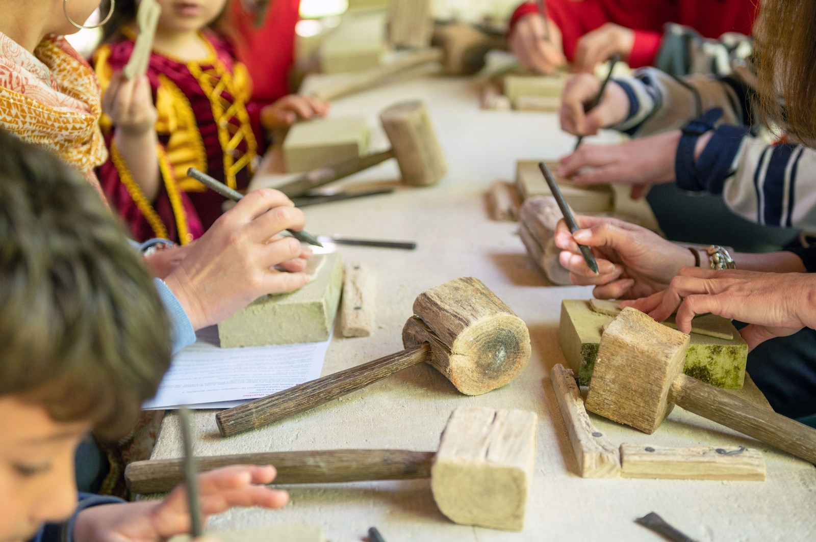 Children and adults sit at a table during a medieval construction workshop, with many hand-built wooden hammers laid out.