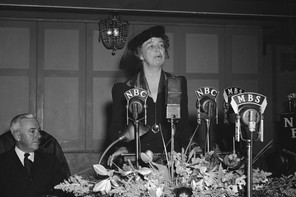 Eleanor Roosevelt stands behind a decorated table, behind several microphones.
