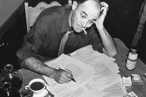 Black-and-white photo of a man smoking a cigarette and filling out forms that are scattered on his desk, with two pots of coffee and one cup of coffee next to the paperwork