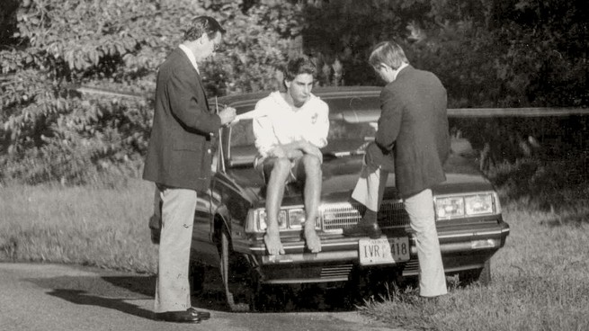 Two men interview a boy sitting on the hood of a car
