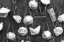 A black-and-white image of various N95 and surgical masks, hanging by their straps from pins on a wooden wall