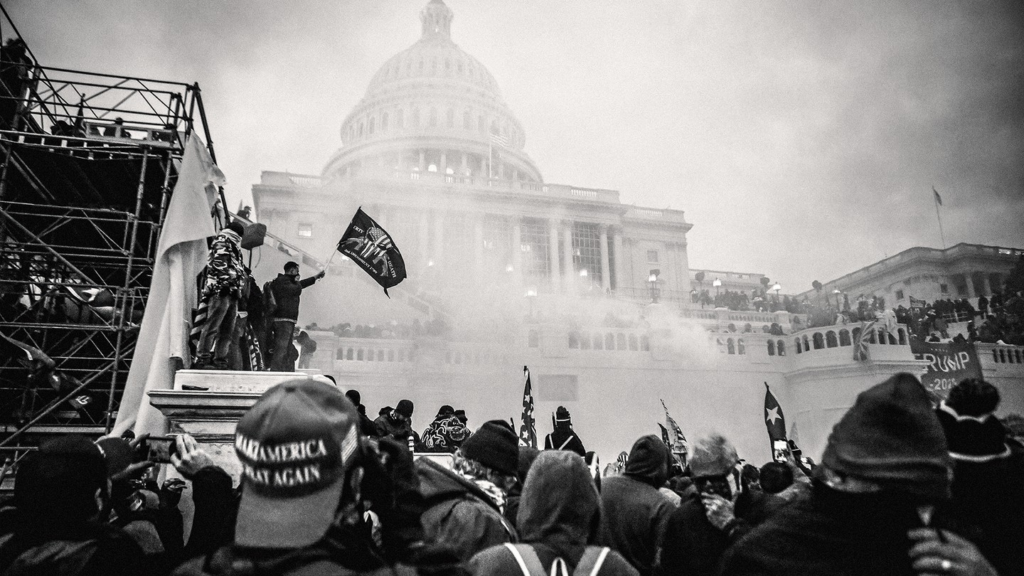 A photo of a crowd with a MAGA hat in the foreground and people standing on scaffolding with a Second Amendment flag and Trump banner in front of the smoke-obscured U.S. Capitol