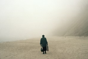 Color photograph of the back of a person in black clothing, carrying bags and standing on a beach