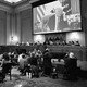 Black-and-white photo of January 6 hearings; photographers kneel on the floor in one part of the room, aiming their cameras at a screen showing Donald Trump on a phone