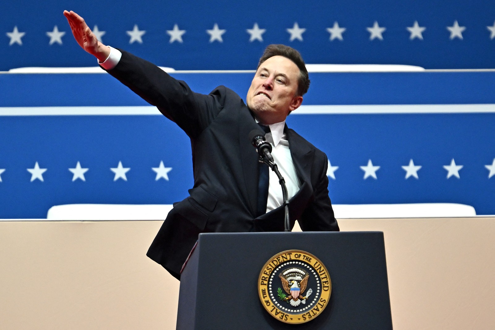 Elon Musk gestures, his right arm held straight out and slightly up, while standing behind a podium with the Presidential seal.