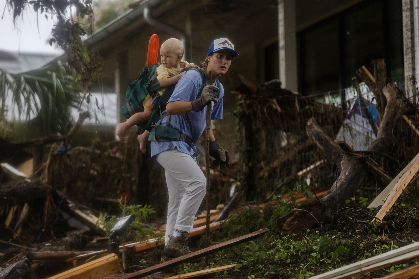 A person searches through flood debris, while carrying a small child in a carrier on their back.