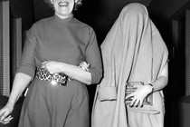 Black and white image of two women walking arm in arm, one with a big smile on her face, the other with a coat over her head, obscuring her face