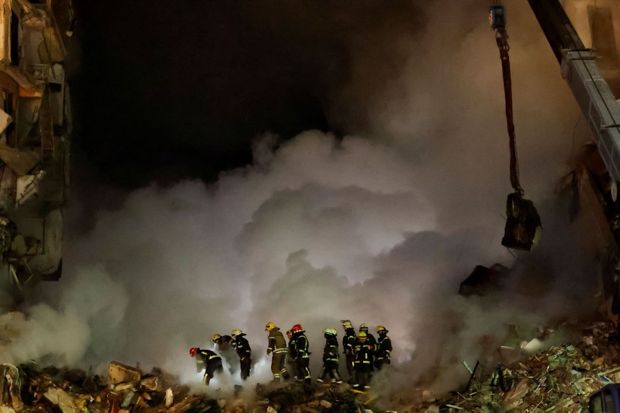 A small crowd of firefighters work on a pile of rubble, beside billowing clouds of smoke.