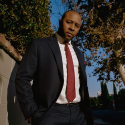 Jarrett in a blue suit outside along a wall with trees in the background