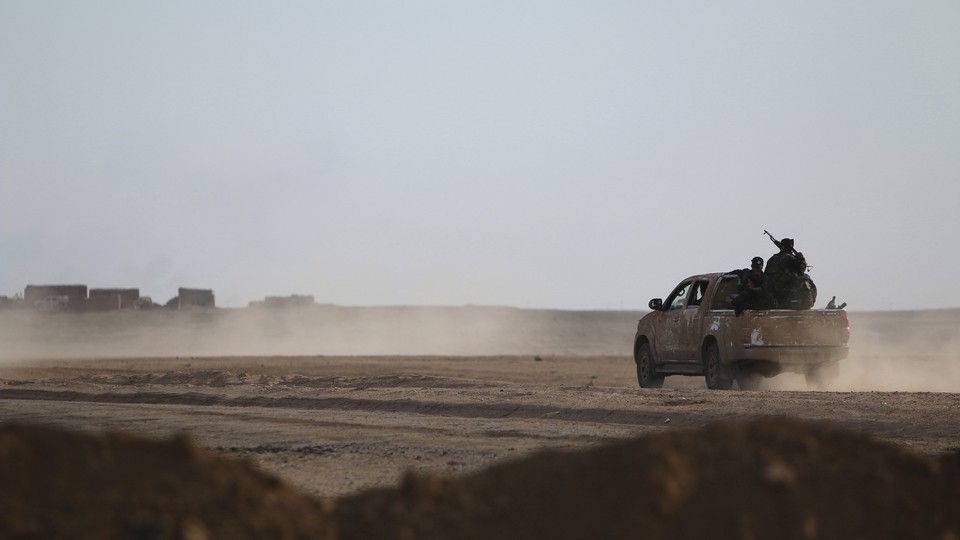 A pickup truck drives on a dusty road toward structures in the distance.