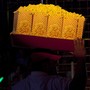 A vendor carries large bags of popcorn on a tray above his head.