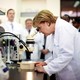 Angela Merkel looks through a microscope while wearing a white lab coat.