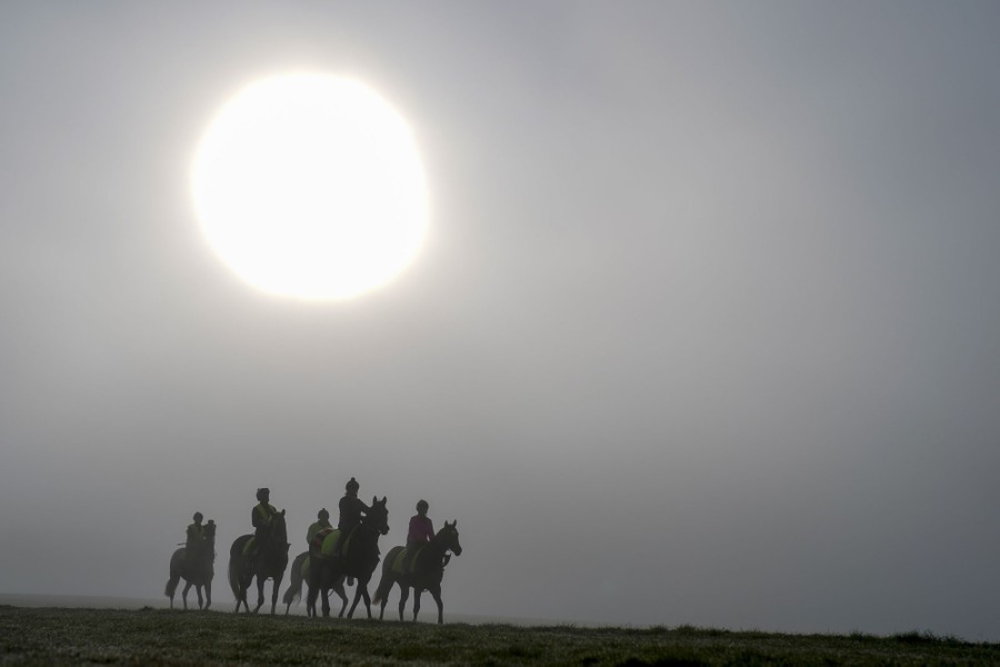 Five riders on horses gallop across a field under a foggy sky and partially obscured sun.