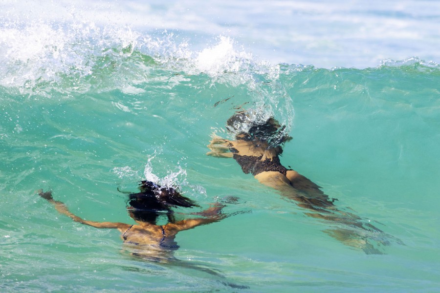 Two women swim under a wave near a beach.