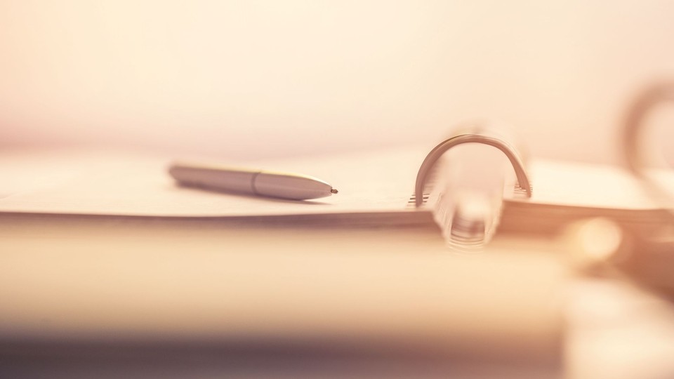 warm, almost sepia-sunlit photo of a white ballpoint pen sitting atop an open spiral notebook