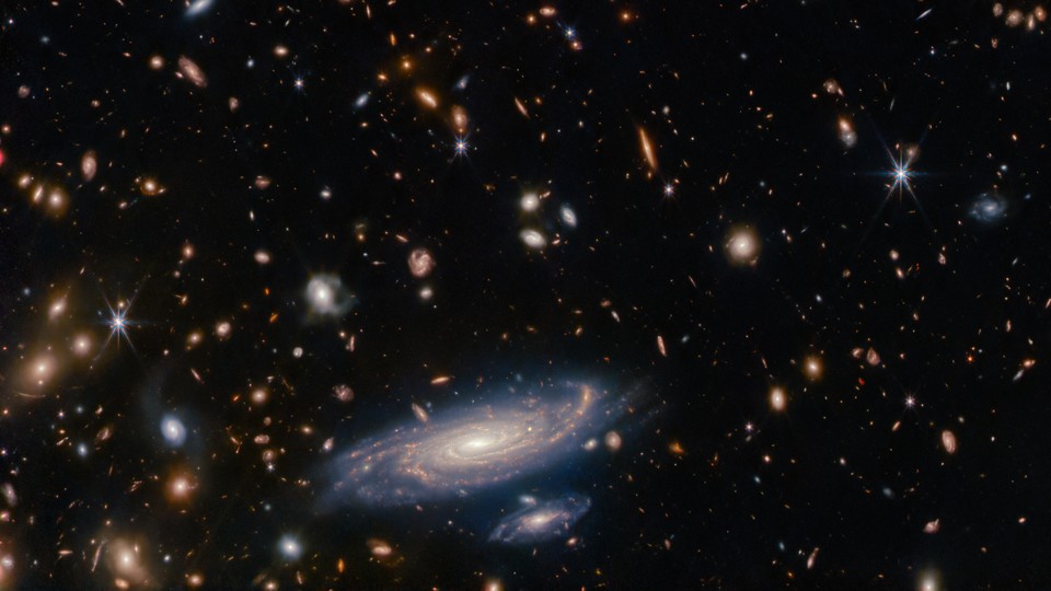 A James Webb Space Telescope picture of a nearby galaxy spiral and sparkling along with many smaller galaxies in the dark background