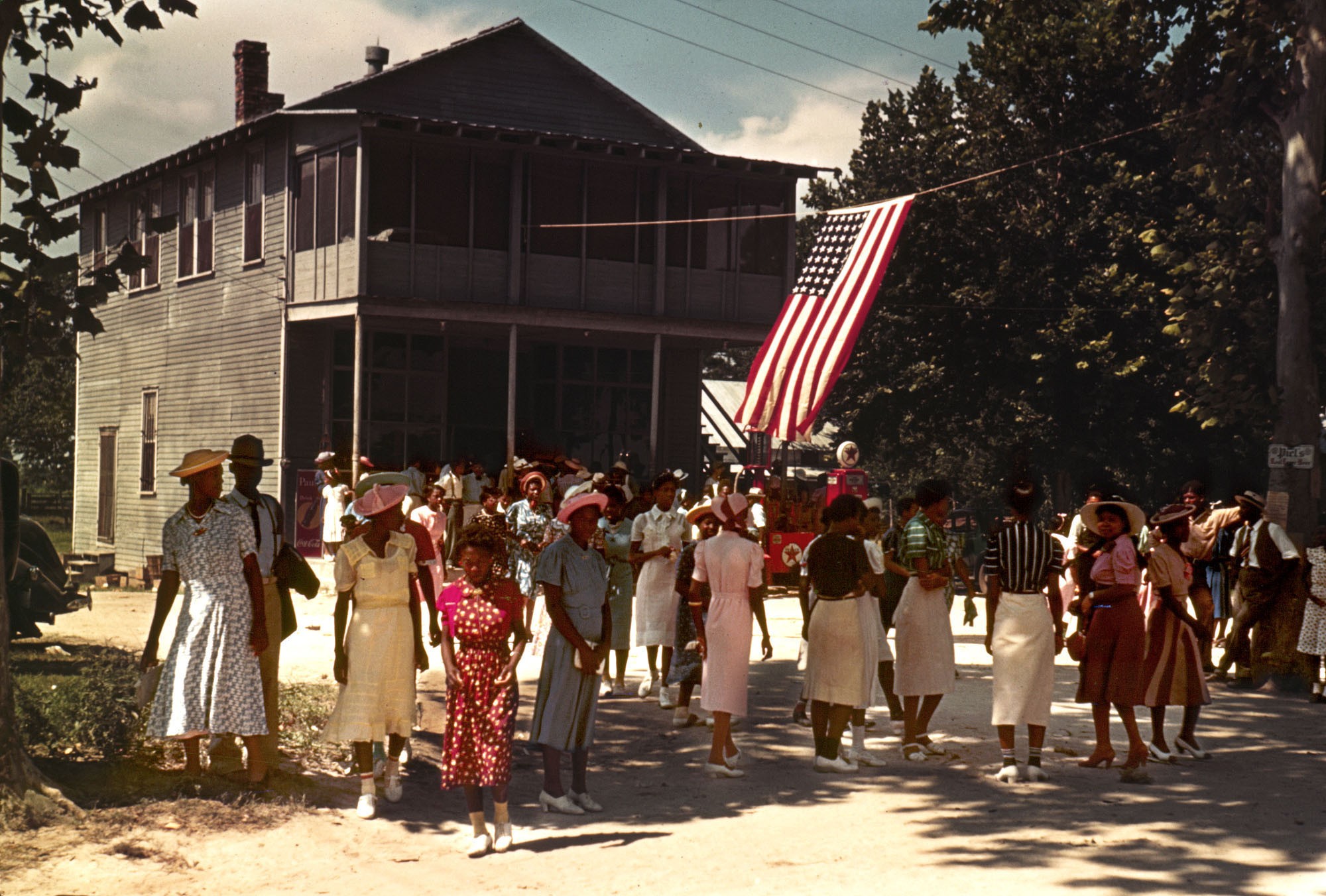 Dozens of well-dressed people stand along a road, beside a Texaco station, on the Fourth of July in 1939.