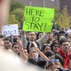 A protester holds a sign that reads "Here to Stay!"