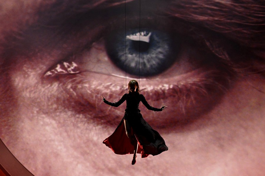 A performer hangs from wires above a stage, in front of a screen showing a large picture of a human eye.