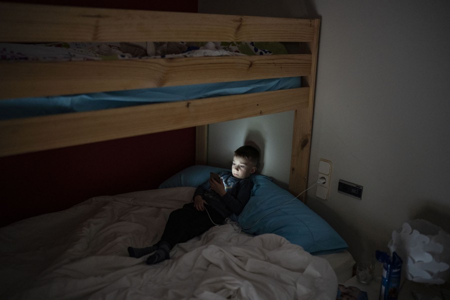 A young child sits in a bed, looking at a cellphone screen.