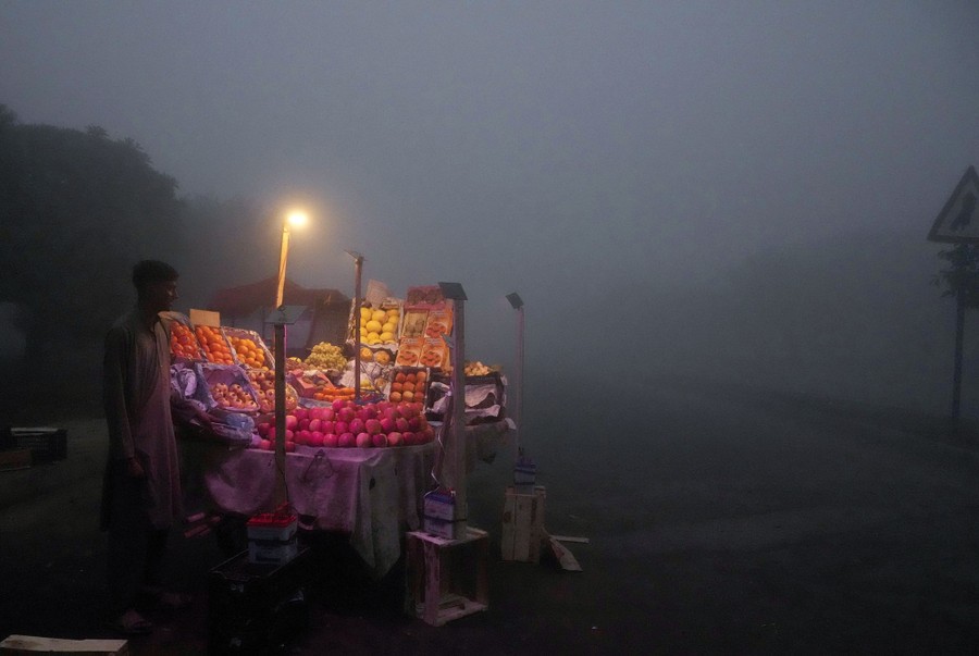A fruit seller stands beside an outdoor display under a dark, smoggy sky.