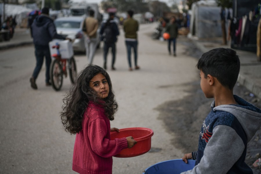 Two children carry plastic tubs in a street as others pass nearby.