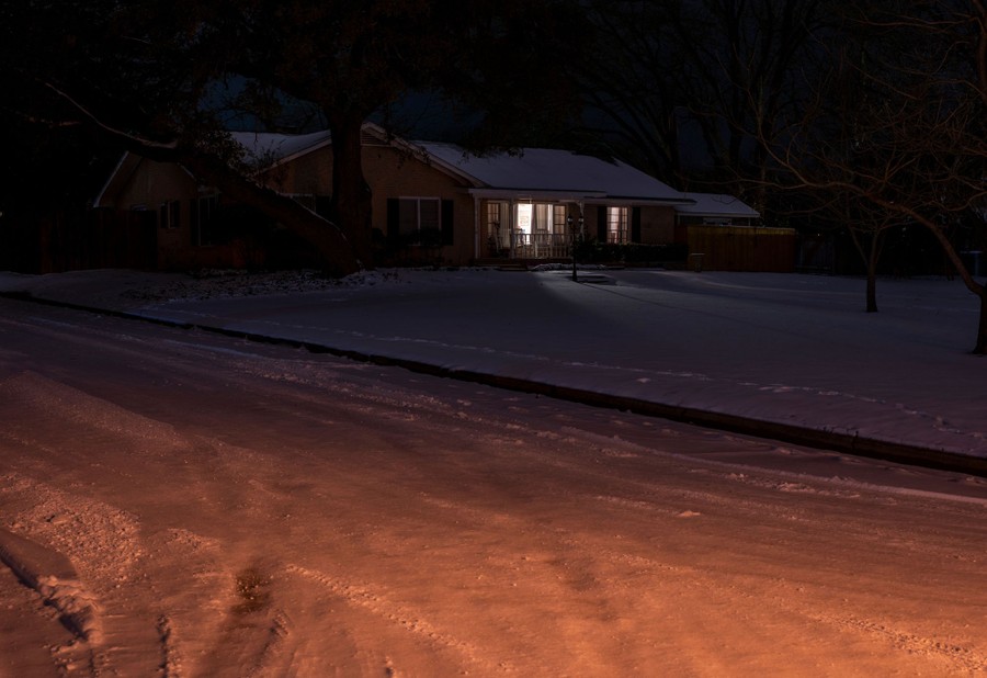 Light comes from inside a house on a dark night, with snow covering the yard and nearby street.