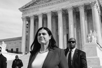 Jena Griswold, wearing a suit jacket and pearls and surrounded by men in dark sunglasses, stands in front of the Supreme Court building.